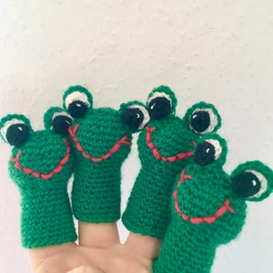 Puede incluir: Cuatro marionetas de dedos de rana de crochet verde con ojos blancos y bocas sonrientes rojas.