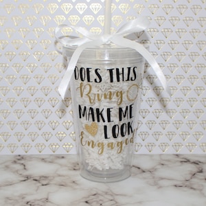 May include: A clear plastic tumbler with a white ribbon and straw. The tumbler has a black and gold glitter design that says "Does this ring make me look engaged?"