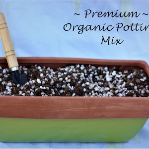 May include: A terracotta planter with a green base filled with a brown and white potting mix. A small gardening trowel with a wooden handle is resting in the mix. The text "~ Premium ~ Organic Potting Mix" is written above the planter.