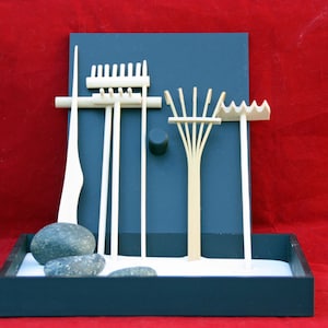 May include: A miniature zen garden with a dark blue frame, white sand, and decorative wooden rakes and stones. The background is a deep red, creating a contrast with the garden's serene elements. The rakes are various shapes.