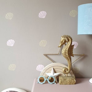 Peut inclure: Une figurine de cheval marin dorée sur un support en bois avec une étoile blanche, entourée d'autocollants muraux en forme de coquillages roses et beiges. La figurine est sur une commode rose avec un coussin en sequins arc-en-ciel et des lunettes de soleil bleues.