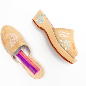 May include: A pair of tan leather mules featuring light blue and orange seahorse and fish illustrations. The interior footbed has a purple label with the text SUSAN BENNIS WARREN EDWARDS and HAND MADE IN ITALY printed in yellow.