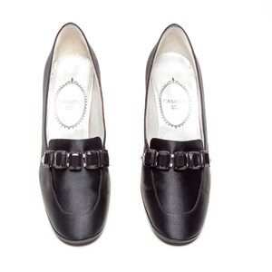 May include: A pair of black satin loafers featuring a row of rectangular black faceted crystals across the vamp. The interior lining is white with the brand name Casadei printed in a decorative oval frame on the heel area of the insole.