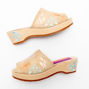 May include: A pair of beige leather slide sandals with a low wedge heel and open toe. The upper features embossed light blue and orange sea life motifs, including fish and seahorses. The insole is purple with a rectangular label.