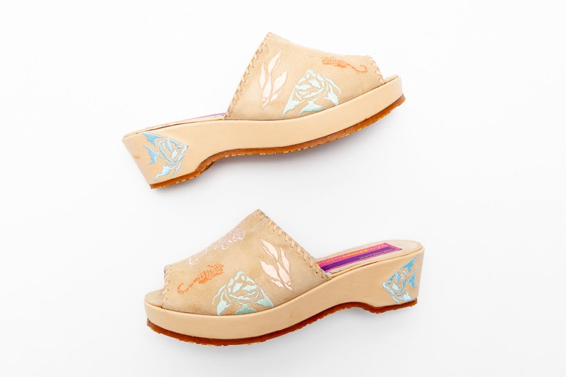 May include: A pair of beige leather slide sandals with a low wedge heel and open toe. The upper features embossed light blue and orange sea life motifs, including fish and seahorses. The insole is purple with a rectangular label.