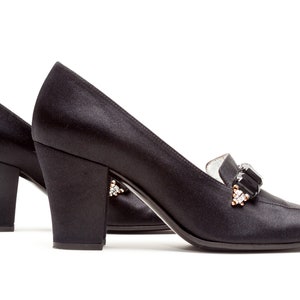 May include: A pair of black satin pumps featuring a block heel and a decorative embellishment on the vamp. The ornament consists of rectangular black stones flanked by smaller clear crystals set in a metallic frame on each shoe.