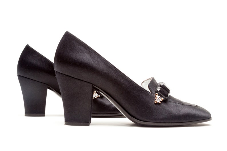 May include: A pair of black satin pumps featuring a block heel and a decorative embellishment on the vamp. The ornament consists of rectangular black stones flanked by smaller clear crystals set in a metallic frame on each shoe.