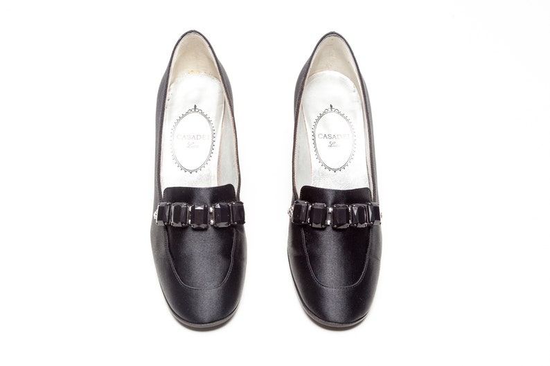 May include: A pair of black satin loafers featuring a row of rectangular black faceted crystals across the vamp. The interior lining is white with the brand name Casadei printed in a decorative oval frame on the heel area of the insole.