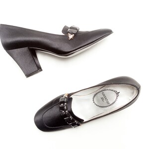 May include: A pair of black satin pumps with a block heel and a square toe. Each shoe features a decorative strap across the vamp adorned with rectangular black crystals and a small cluster of clear stones. The insole is silver with Casadei logo.