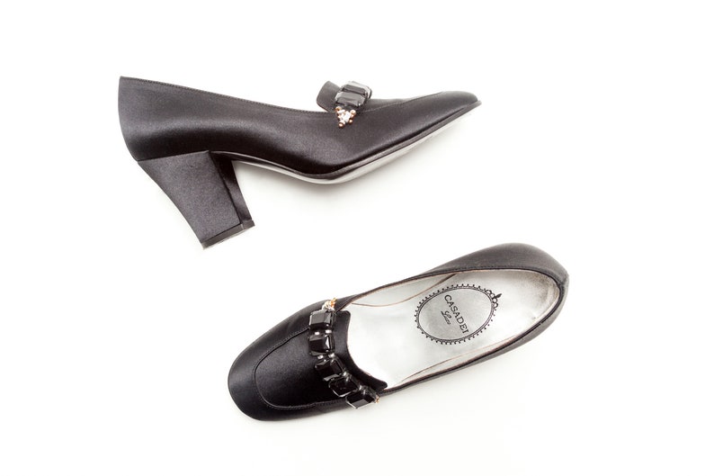 May include: A pair of black satin pumps with a block heel and a square toe. Each shoe features a decorative strap across the vamp adorned with rectangular black crystals and a small cluster of clear stones. The insole is silver with Casadei logo.