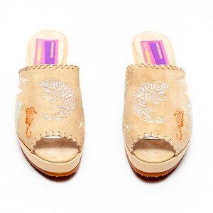 May include: A pair of beige suede open-toe mules featuring embroidered white fish and orange seahorse motifs. The shoes have a thick platform sole, a tan textured outsole, and a rectangular purple and yellow label on the inner heel.