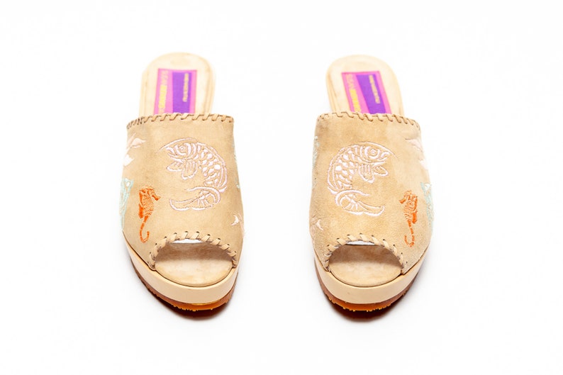 May include: A pair of beige suede open-toe mules featuring embroidered white fish and orange seahorse motifs. The shoes have a thick platform sole, a tan textured outsole, and a rectangular purple and yellow label on the inner heel.