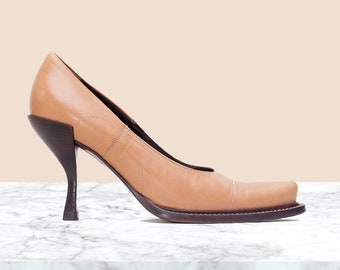 Puede incluir: Zapato de tacón de cuero color canela con un tacón angular único y una puntera cuadrada. El zapato presenta un diseño cosido en la parte superior y una suela marrón oscuro. El tacón mide unos 10 cm de alto.