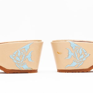 May include: A pair of beige wedge sandals featuring a light blue embroidered angelfish design on the side of each heel. The shoes have a smooth, light-colored finish and a brown sole, showcasing a detailed fish motif on the outer heel area.