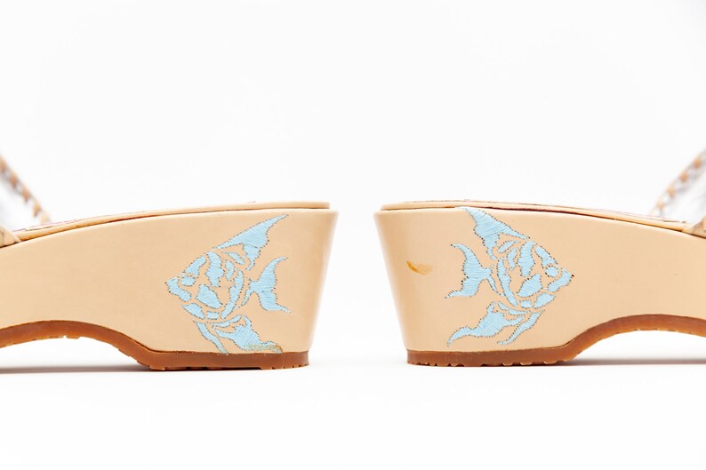 May include: A pair of beige wedge sandals featuring a light blue embroidered angelfish design on the side of each heel. The shoes have a smooth, light-colored finish and a brown sole, showcasing a detailed fish motif on the outer heel area.