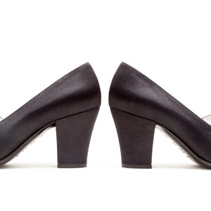 May include: A pair of black satin pumps with a block heel and a decorative rectangular black stone accent featuring small clear crystals at the base. The shoes have a smooth finish and a structured silhouette, presented side by side.