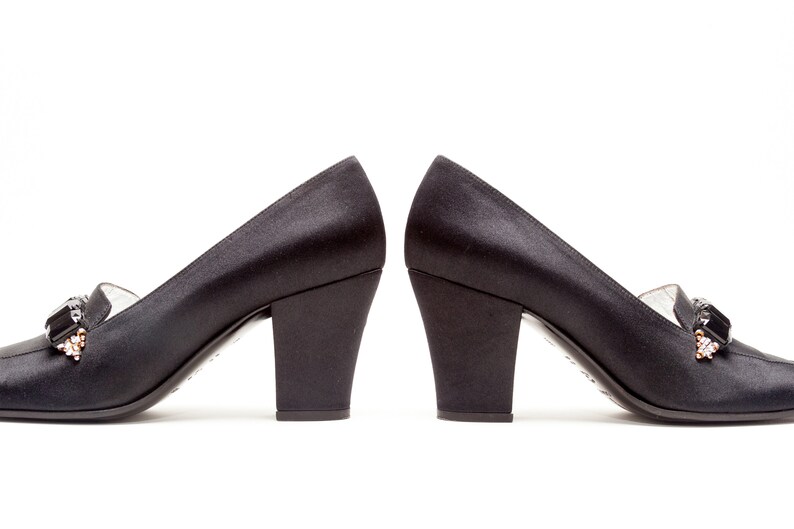 May include: A pair of black satin pumps with a block heel and a decorative rectangular black stone accent featuring small clear crystals at the base. The shoes have a smooth finish and a structured silhouette, presented side by side.