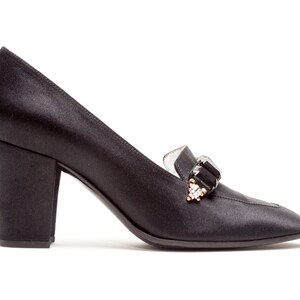 May include: A black satin pump with a block heel and a rounded toe. The shoe features a decorative embellishment on the vamp consisting of a large rectangular black stone flanked by smaller clear crystals set in a gold-tone metal base.