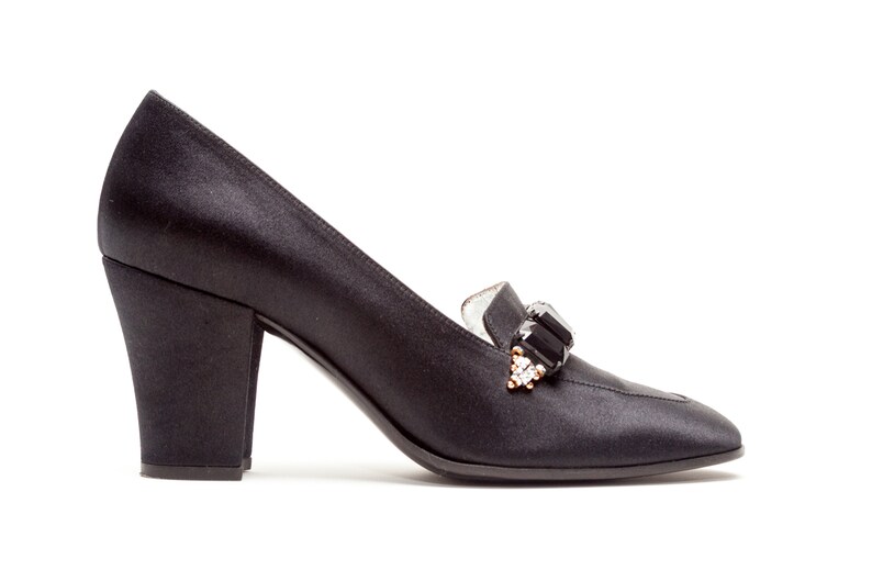 May include: A black satin pump with a block heel and a rounded toe. The shoe features a decorative embellishment on the vamp consisting of a large rectangular black stone flanked by smaller clear crystals set in a gold-tone metal base.