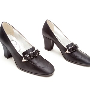 May include: A pair of black satin pumps featuring a block heel and a rounded toe. Each shoe is adorned with a decorative strap across the vamp, embellished with a row of large, rectangular black crystals and small clear accent stones.