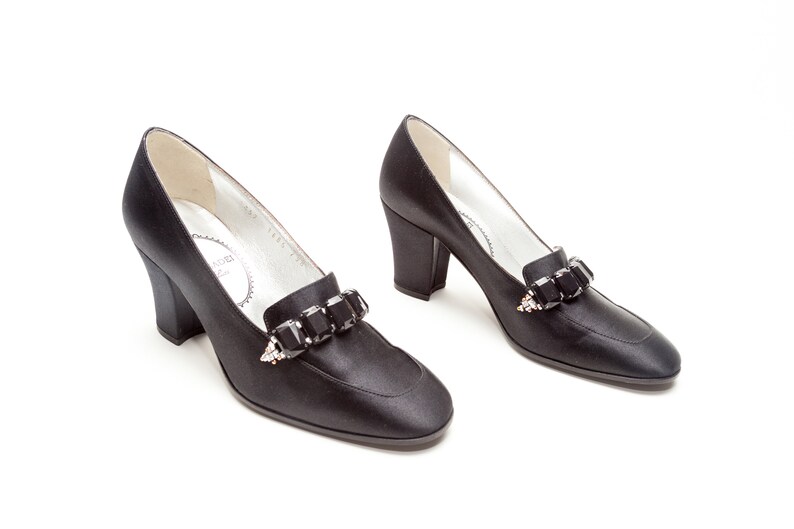 May include: A pair of black satin pumps featuring a block heel and a rounded toe. Each shoe is adorned with a decorative strap across the vamp, embellished with a row of large, rectangular black crystals and small clear accent stones.