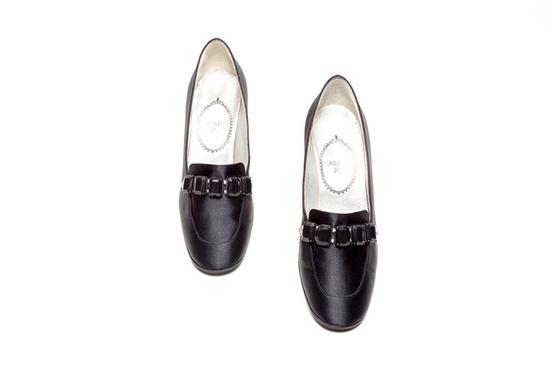 May include: A pair of black satin loafers featuring a rounded toe and a decorative strap across the vamp adorned with rectangular black crystals. The interior lining is white with the text Casadei printed inside an oval frame on the insole.