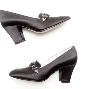 May include: A pair of black satin pumps featuring a block heel and a decorative strap across the vamp adorned with rectangular black crystals and small clear stones. The shoes have a rounded toe and a smooth, polished finish.