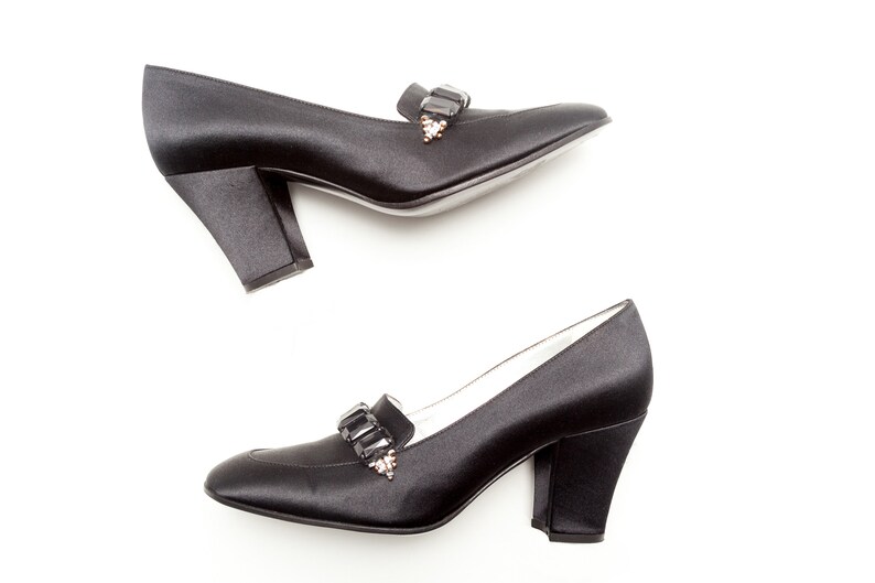 May include: A pair of black satin pumps featuring a block heel and a decorative strap across the vamp adorned with rectangular black crystals and small clear stones. The shoes have a rounded toe and a smooth, polished finish.