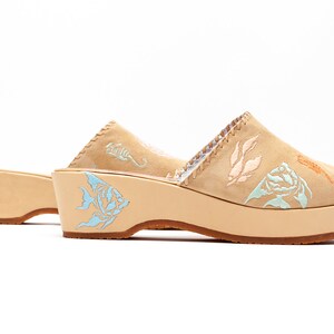 May include: A pair of tan suede platform mules featuring light blue, peach, and orange embroidered marine life motifs including fish, seahorses, and seaweed. The shoes have a thick, light-colored wooden platform sole and a brown rubber outsole.