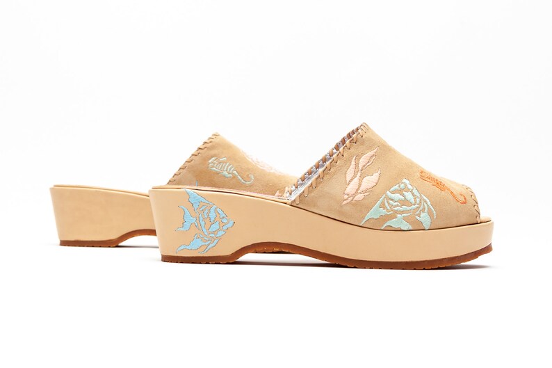 May include: A pair of tan suede platform mules featuring light blue, peach, and orange embroidered marine life motifs including fish, seahorses, and seaweed. The shoes have a thick, light-colored wooden platform sole and a brown rubber outsole.