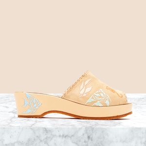 May include: A beige suede mule with a wedge heel, featuring light blue and white floral embroidery on the upper and a matching floral design on the side of the wooden heel. The shoe has a peep-toe opening and visible stitching along the edges.