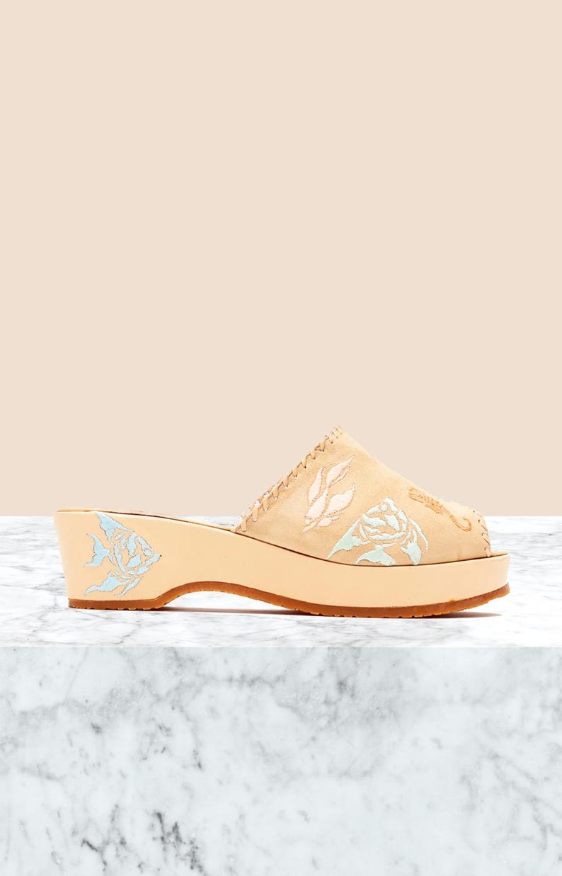May include: A beige suede mule with a wedge heel, featuring light blue and white floral embroidery on the upper and a matching floral design on the side of the wooden heel. The shoe has a peep-toe opening and visible stitching along the edges.