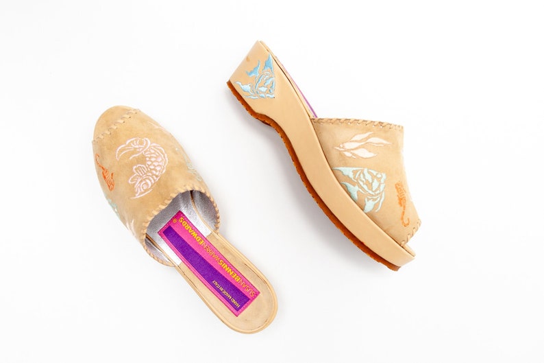 May include: A pair of tan leather mules featuring light blue and orange seahorse and fish illustrations. The interior footbed has a purple label with the text SUSAN BENNIS WARREN EDWARDS and HAND MADE IN ITALY printed in yellow.