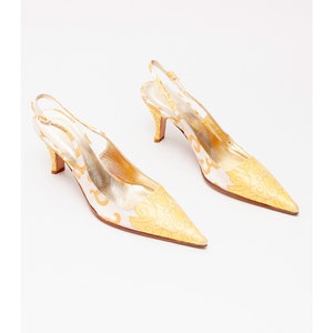 Puede incluir: Un par de tacones blancos de tiras con un estampado floral dorado. Los tacones tienen una punta puntiaguda y una tira fina que va alrededor del tobillo.