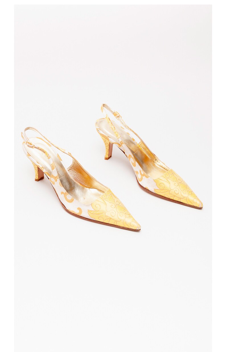 Puede incluir: Un par de tacones blancos de tiras con un estampado floral dorado. Los tacones tienen una punta puntiaguda y una tira fina que va alrededor del tobillo.