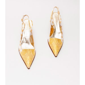 Puede incluir: Un par de tacones blancos y dorados de slingback con punta puntiaguda y un delicado estampado floral. Los tacones est&aacute;n hechos de un material satinado y tienen una hebilla dorada en la correa del tobillo.