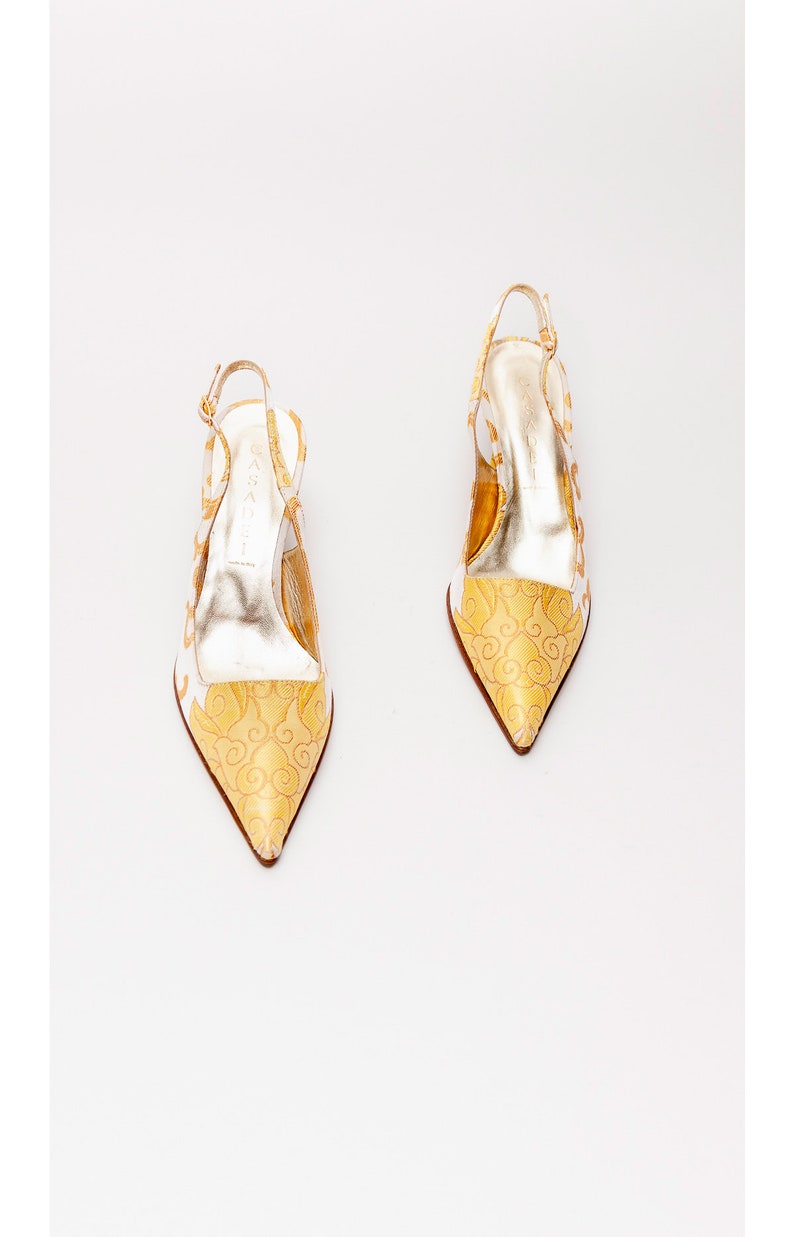 Puede incluir: Un par de tacones blancos y dorados de slingback con punta puntiaguda y un delicado estampado floral. Los tacones est&aacute;n hechos de un material satinado y tienen una hebilla dorada en la correa del tobillo.