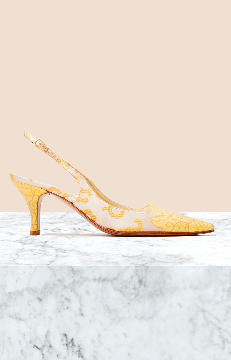 Puede incluir: Un par de tacones amarillos y blancos con tira en el tobillo y un estampado floral. Los tacones est&aacute;n sobre una superficie de m&aacute;rmol blanco.