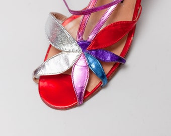 May include: A pair of red, silver, purple, and blue leather sandals with a flower design. The sandals have a thin ankle strap and a small heel.