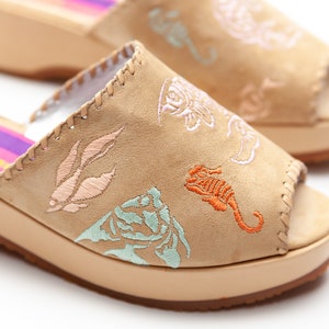 May include: A pair of beige suede wedge sandals featuring open toes and embroidered marine life motifs including a seahorse, shells, and coral in shades of orange, light blue, and pink. The inner sole displays a purple and pink striped pattern.