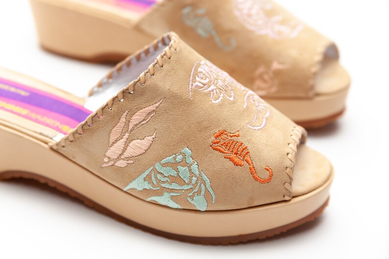 May include: A pair of beige suede wedge sandals featuring open toes and embroidered marine life motifs including a seahorse, shells, and coral in shades of orange, light blue, and pink. The inner sole displays a purple and pink striped pattern.
