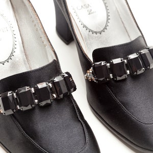 May include: A pair of black satin pumps featuring a block heel and a decorative strap across the vamp adorned with five large, rectangular, faceted black crystals. The interior silver-toned insole displays the text CASADEI.