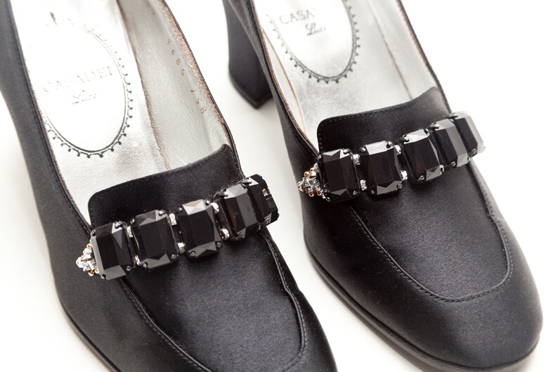 May include: A pair of black satin pumps featuring a block heel and a decorative strap across the vamp adorned with five large, rectangular, faceted black crystals. The interior silver-toned insole displays the text CASADEI.
