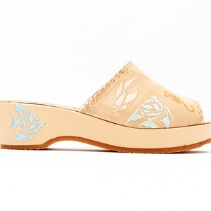 May include: A beige suede mule with a platform wedge heel. The upper features embroidered light blue and white leaf and floral patterns. A light blue fish design is embroidered on the side of the heel, and a seahorse is on the toe.