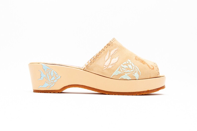 May include: A beige suede mule with a platform wedge heel. The upper features embroidered light blue and white leaf and floral patterns. A light blue fish design is embroidered on the side of the heel, and a seahorse is on the toe.