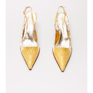 Puede incluir: Un par de tacones blancos de tiras con un dise&ntilde;o estampado en dorado. Los tacones son de punta y tienen una tira fina alrededor del tobillo.
