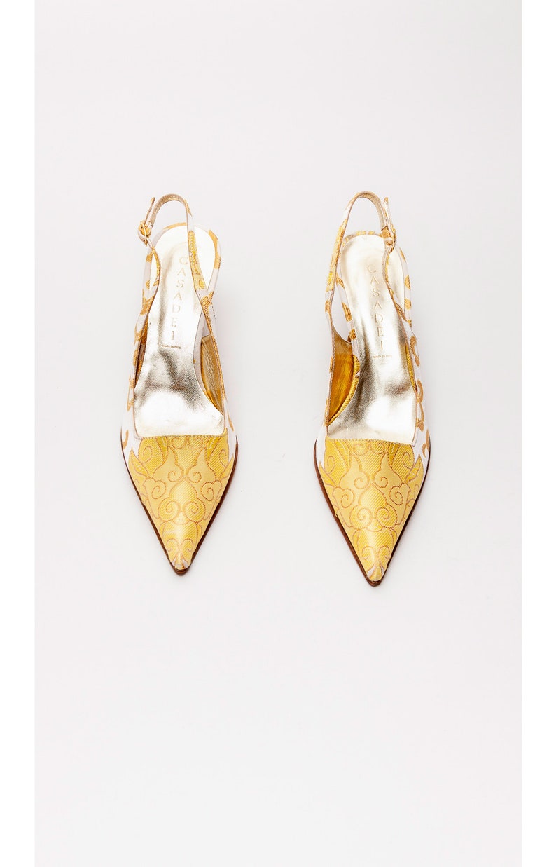 Puede incluir: Un par de tacones blancos de tiras con un dise&ntilde;o estampado en dorado. Los tacones son de punta y tienen una tira fina alrededor del tobillo.
