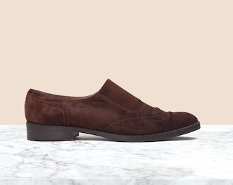 May include: A brown suede slip-on shoe with a low heel. The shoe features decorative perforations along the edges and a smooth, dark sole. The shoe is displayed on a marble surface.