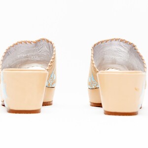 May include: A pair of beige wedge sandals featuring a light blue floral pattern on the sides, a silver interior lining, and a thick beige heel. The shoes are shown from the back, highlighting the solid heel construction and the stitching.