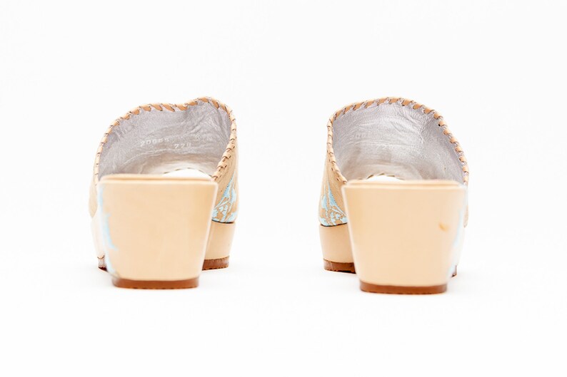 May include: A pair of beige wedge sandals featuring a light blue floral pattern on the sides, a silver interior lining, and a thick beige heel. The shoes are shown from the back, highlighting the solid heel construction and the stitching.